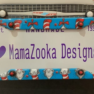 May include: A blue and white license plate frame with a cat in a hat design. The frame has red and white accents and says "Est 1997 Handmade MamaZoooka Designs".