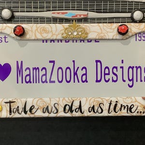 May include: A white and gold decorative license plate frame with a rose design. It features the text "Est 1997", "MamaZooka Designs", and "Tale as old as time..." with a purple heart and a gold crown.