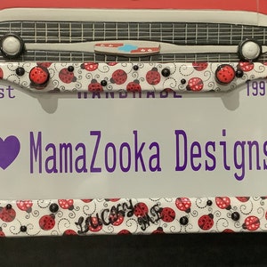 May include: White metal license plate frame with a ladybug design. The frame has a purple heart and the text "MamaZooka Designs" in purple. The frame also has the text "Est 1997".