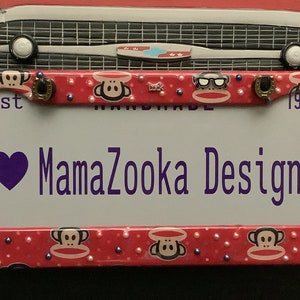 May include: A red license plate frame with a repeating monkey pattern and the text "MamaZooka Designs." The frame has a white background with a purple heart. The frame is decorated with small pearls and blue beads.