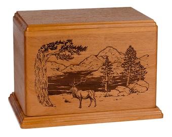 May include: A wooden cremation urn with an engraved image of a mountain scene with a lake, trees, and an elk. The urn is a light brown color.