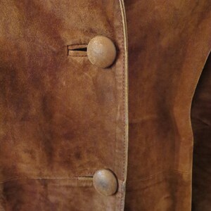May include: Close-up of a brown suede jacket with two large, round, brown buttons. The jacket has a seam down the front and a light brown trim.