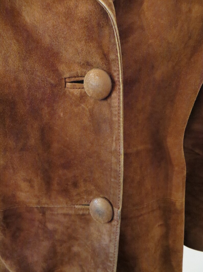 May include: Close-up of a brown suede jacket with two large, round, brown buttons. The jacket has a seam down the front and a light brown trim.