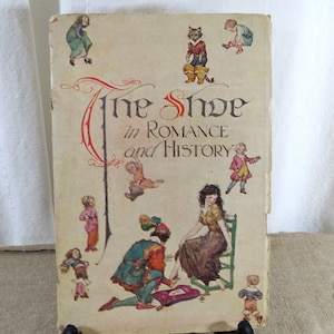May include: A vintage book cover with the title "The Shoe in Romance and History". The cover features a colorful illustration of a woman sitting on a chair while a man kneels and ties her shoe. There are other small illustrations of people and animals around the main illustration.