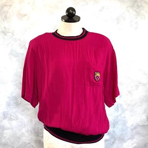 May include: A magenta short-sleeved shirt with a black and dark red trim around the neckline and hem. The shirt has a small pocket on the chest with a gold and black emblem. The shirt is displayed on a mannequin.