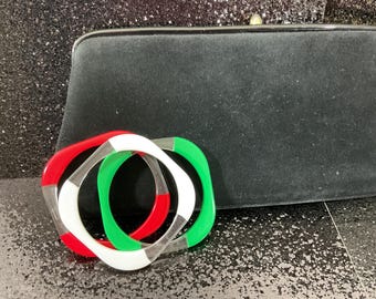 May include: A black clutch bag with a rectangular shape and a clasp closure. Three square-shaped bangles in red, white, and green are in front of the bag. The bangles have a geometric design.