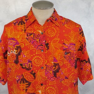 May include: A red short-sleeved button-down shirt with a colorful geometric and floral pattern. The shirt has a button-up collar and a pocket on the left side.