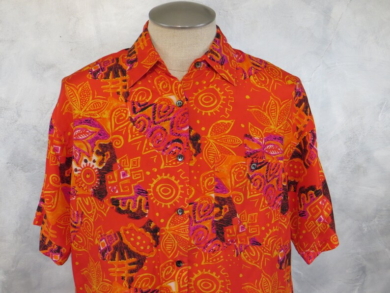 May include: A red short-sleeved button-down shirt with a colorful geometric and floral pattern. The shirt has a button-up collar and a pocket on the left side.