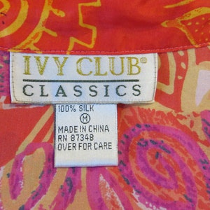 May include: A white tag with the text "IVY CLUB CLASSICS" in black lettering. The tag also says "100% SILK" and "MADE IN CHINA RN 87348 OVER FOR CARE". The tag is attached to a red and orange fabric with a floral pattern.