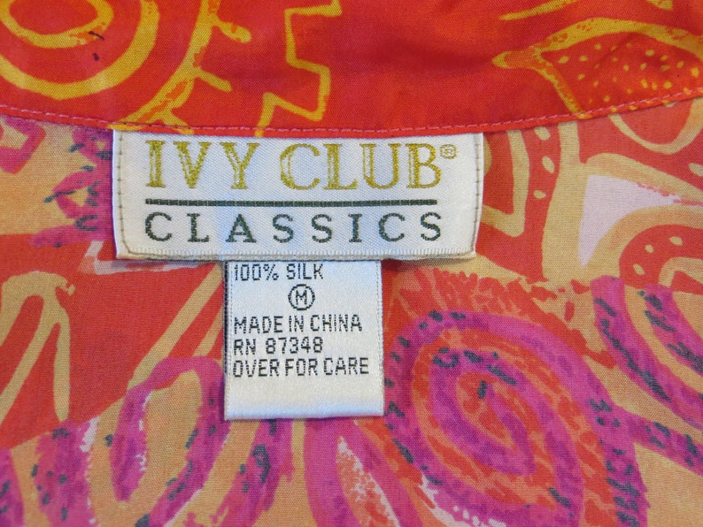 May include: A white tag with the text "IVY CLUB CLASSICS" in black lettering. The tag also says "100% SILK" and "MADE IN CHINA RN 87348 OVER FOR CARE". The tag is attached to a red and orange fabric with a floral pattern.