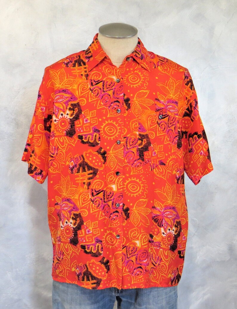May include: A short-sleeved button-down shirt with a bright orange background and a colorful abstract pattern. The pattern features pink, orange, yellow, and black geometric shapes and designs.