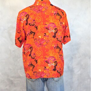 May include: A short-sleeved button-up shirt with a bright orange background and a colorful abstract pattern. The pattern features pink, orange, and black geometric shapes and designs.