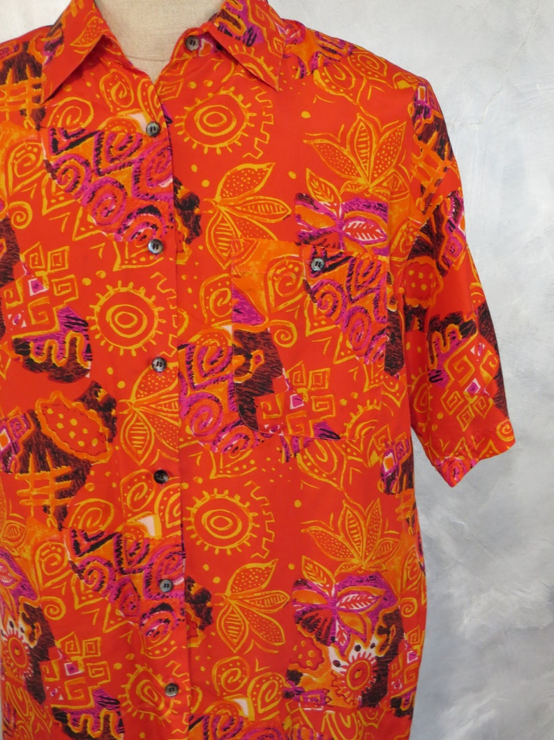 May include: A short-sleeved button-down shirt with a vibrant orange, yellow, and pink geometric pattern. The shirt has a collar and a pocket on the left side.