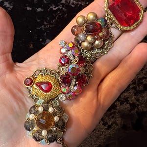 May include: A collection of vintage brooches and pendants in various shapes and sizes. The jewelry features red, gold, and iridescent rhinestones, faux pearls, and faceted glass beads. The pieces are displayed on a hand.
