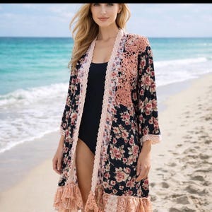 以下が含まれることがあります： 黒を基調とした花柄の着物で、ピンクと赤の花が特徴です。着物には、ピーチ色のかぎ針編みのパネルとトリム、フリル付きの裾が付いています。ビーチで黒い水着の上に着用しています。