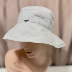 May include: A solid off-white bucket hat with a wide, downward-sloping brim and a small, dark oval-shaped metal emblem centered on the front band. The hat is constructed from a textured fabric and features visible stitching along the brim and crown.