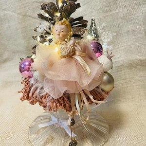 Puede incluir: Adorno de árbol de Navidad hecho a mano con una muñeca de porcelana con un vestido de tul rosa. El árbol está hecho de piñas, adornos y luces de hadas, coronado con un broche vintage. La base es un pedestal de vidrio transparente.