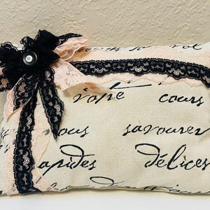 May include: Rectangular decorative pillow with a cream-colored fabric base. The pillow features black script writing, black lace trim, and a black bow with a pearl button. Pink lace accents the bow and trim.