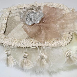 May include: An ornate, cream-colored decorative piece with a clear, faceted glass knob at the center. It features a beige bow, a crocheted border, and dangling tassels. The base has a fluffy, white texture.