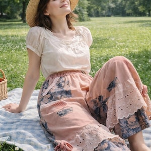 Puede incluir: Una persona con sombrero de paja y blusa crema con falda floral, sentada en una manta a cuadros azul y blanco en un parque. Una cesta de picnic y un plato de fresas están cerca.