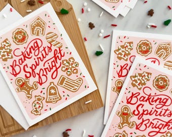 May include: Pink holiday greeting cards with a gingerbread cookie theme. The cards feature a variety of gingerbread cookies, including a gingerbread man, a gingerbread house, a gingerbread tree, and a gingerbread stocking. The cards also feature the text "Baking Spirits Bright".