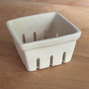 May include: A square, off-white ceramic berry basket with a matte finish. The container features a thick, rounded rim and vertical rectangular cutouts along all four sides, mimicking the appearance of a traditional wooden produce crate.