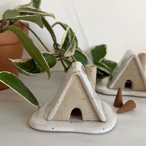 以下が含まれることがあります： 小さな煙突のある家型の白いセラミック製のお香立て。お香立ては、斑点のある仕上げの白いセラミック製の台座の上に置かれています。2つのコーン型のお香が台座の上に置かれています。