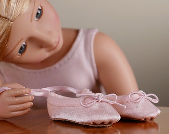 May include: A pair of pink satin ballet shoes with satin ribbons. The shoes are on a wooden surface and a doll is tying the ribbon on one of the shoes.