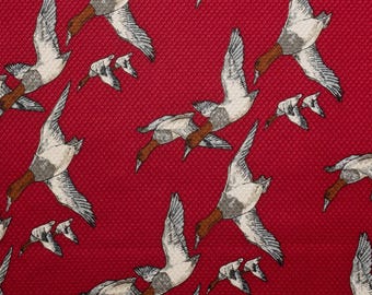 May include: Red silk twill fabric with a repeating pattern of Canvasback ducks in flight. The ducks are depicted in shades of white, gray, and brown against a vibrant red background. The fabric has a textured appearance. The text on the fabric reads "#227A Red Canvasback Ducks 100% Silk Twill".