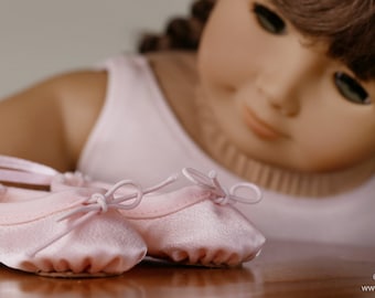 May include: A pair of pink satin ballet shoes with a bow on each shoe. The shoes are on a wooden surface and a doll is looking down at them.