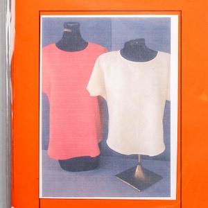 May include: An orange package with the text "BASIC SHELL" and a signature. Inside, a photo shows two t-shirts on mannequins: one coral and one white. The package is sealed in a clear plastic bag.