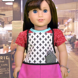 May include: A doll wearing a red and white patterned shirt, a black and white apron with a fleur-de-lis design, a pink skirt, and a red and white bracelet. The doll is standing in front of a cafe.