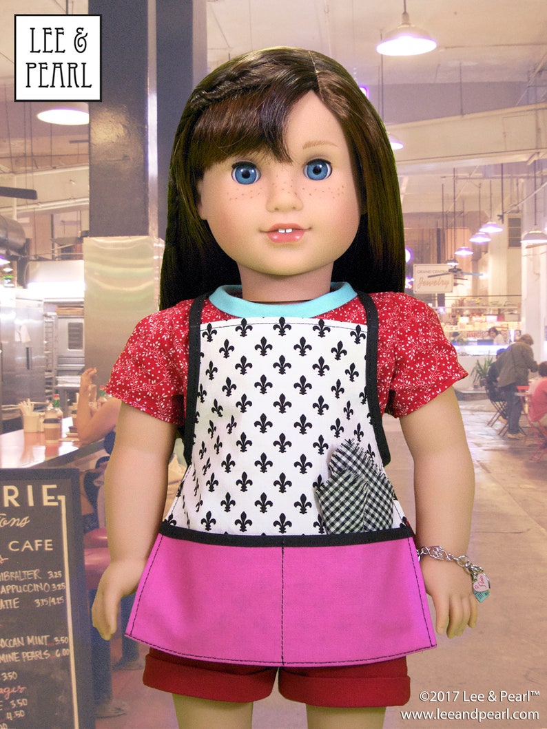 May include: A doll wearing a red and white patterned shirt, a black and white apron with a fleur-de-lis design, a pink skirt, and a red and white bracelet. The doll is standing in front of a cafe.