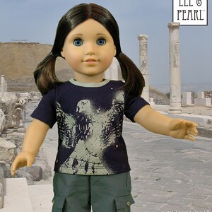 May include: A doll wearing a dark blue t-shirt with a white graphic of a bird on it. The doll is also wearing green cargo shorts. The doll has brown hair and blue eyes. The doll is standing in front of a stone wall.