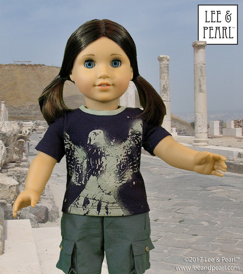 May include: A doll wearing a dark blue t-shirt with a white graphic of a bird on it. The doll is also wearing green cargo shorts. The doll has brown hair and blue eyes. The doll is standing in front of a stone wall.
