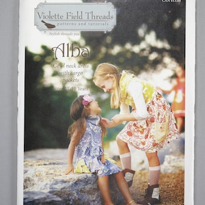 May include: A sewing pattern for a cowl neck dress with cargo pockets for children. The pattern is called "Alba" and is from Violette Field Threads. The pattern is for sizes 2T-10 years.