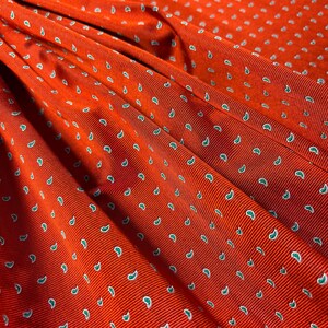 May include: A vibrant orange fabric featuring a repeating pattern of small, white, teardrop-shaped paisley motifs. The material has a distinct ribbed texture with visible diagonal lines running across the surface, creating depth and dimension.