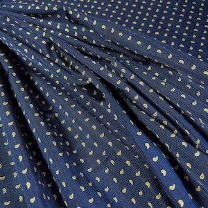 May include: A dark blue fabric featuring a repeating pattern of small, yellow, teardrop-shaped paisley motifs. The material has a fine, ribbed texture and is draped in soft folds, creating a series of shadows and highlights across the surface.