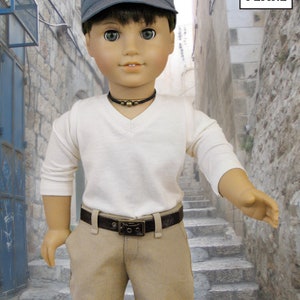 May include: A doll wearing a white long-sleeved shirt, a brown belt, khaki pants, and a dark blue cap. The doll is standing in a narrow alleyway with stone walls and steps.