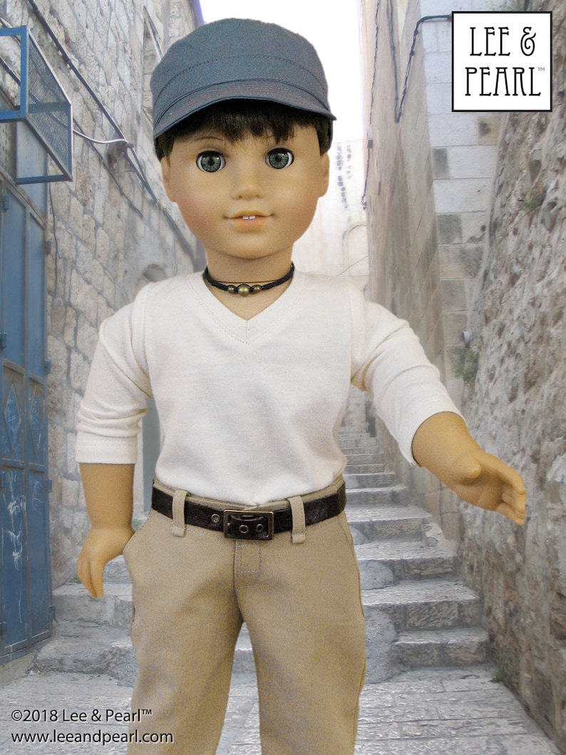 May include: A doll wearing a white long-sleeved shirt, a brown belt, khaki pants, and a dark blue cap. The doll is standing in a narrow alleyway with stone walls and steps.
