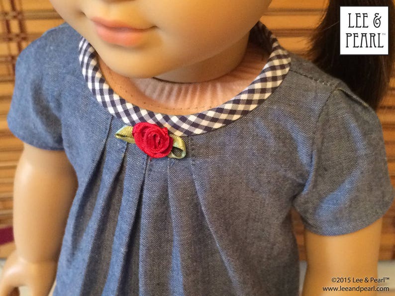 May include: A blue denim dress with a white and black gingham collar and a red fabric flower accent. The dress has a pleated bodice and short sleeves.