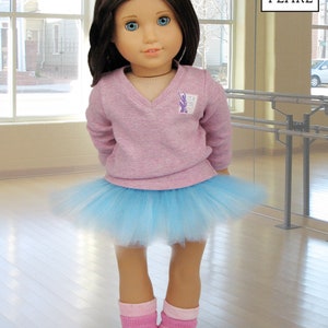May include: A young girl doll wearing a pink sweater with a purple logo that says "Lee & Pearl", a light blue tulle tutu, pink socks, and pink ballet shoes. The doll is standing on a wooden floor in front of a mirror.