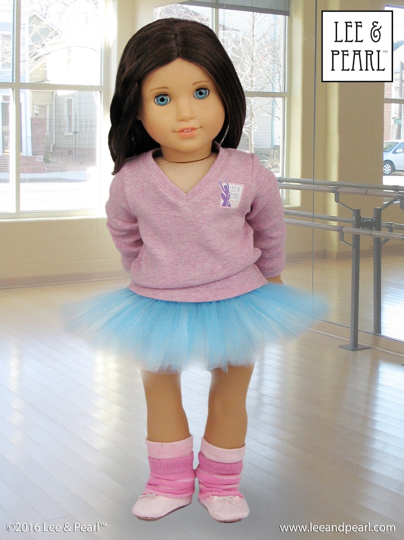 May include: A young girl doll wearing a pink sweater with a purple logo that says "Lee & Pearl", a light blue tulle tutu, pink socks, and pink ballet shoes. The doll is standing on a wooden floor in front of a mirror.