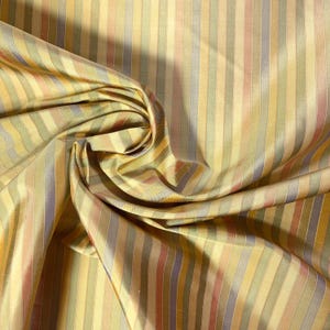 May include: A piece of fabric featuring a vertical striped pattern in shades of gold, sage green, muted purple, and soft orange. The material has a subtle sheen and is draped to create soft folds, highlighting the repeating linear design.