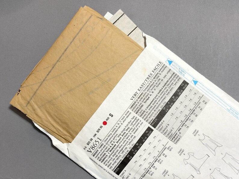 May include: A sewing pattern packet with a tan paper pattern peeking out. The packet is white with black text and diagrams, including the words "VERY EASY/TR&Egrave;S FACILE" and "V8651". The pattern is for a tunic.