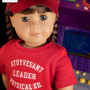 May include: A red t-shirt with white text that reads "Stuyvesant Leader Physical Ed." The shirt is being worn by a doll with brown hair and green eyes. The doll is wearing a red baseball cap.