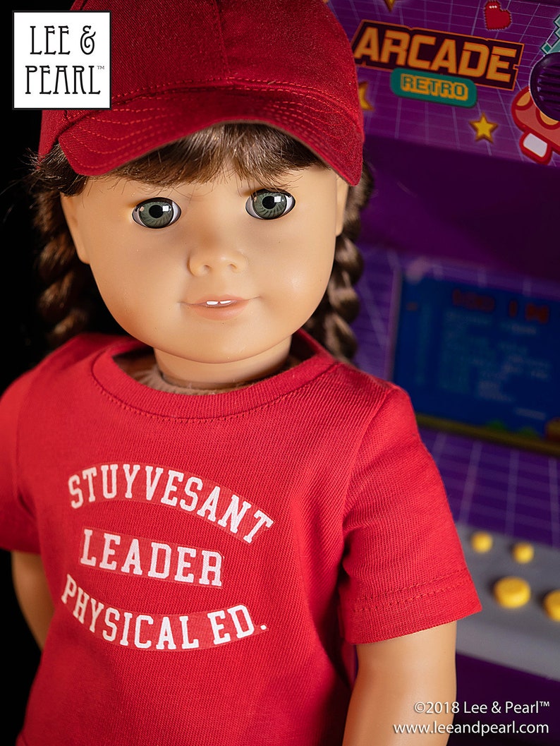 May include: A red t-shirt with white text that reads "Stuyvesant Leader Physical Ed." The shirt is being worn by a doll with brown hair and green eyes. The doll is wearing a red baseball cap.