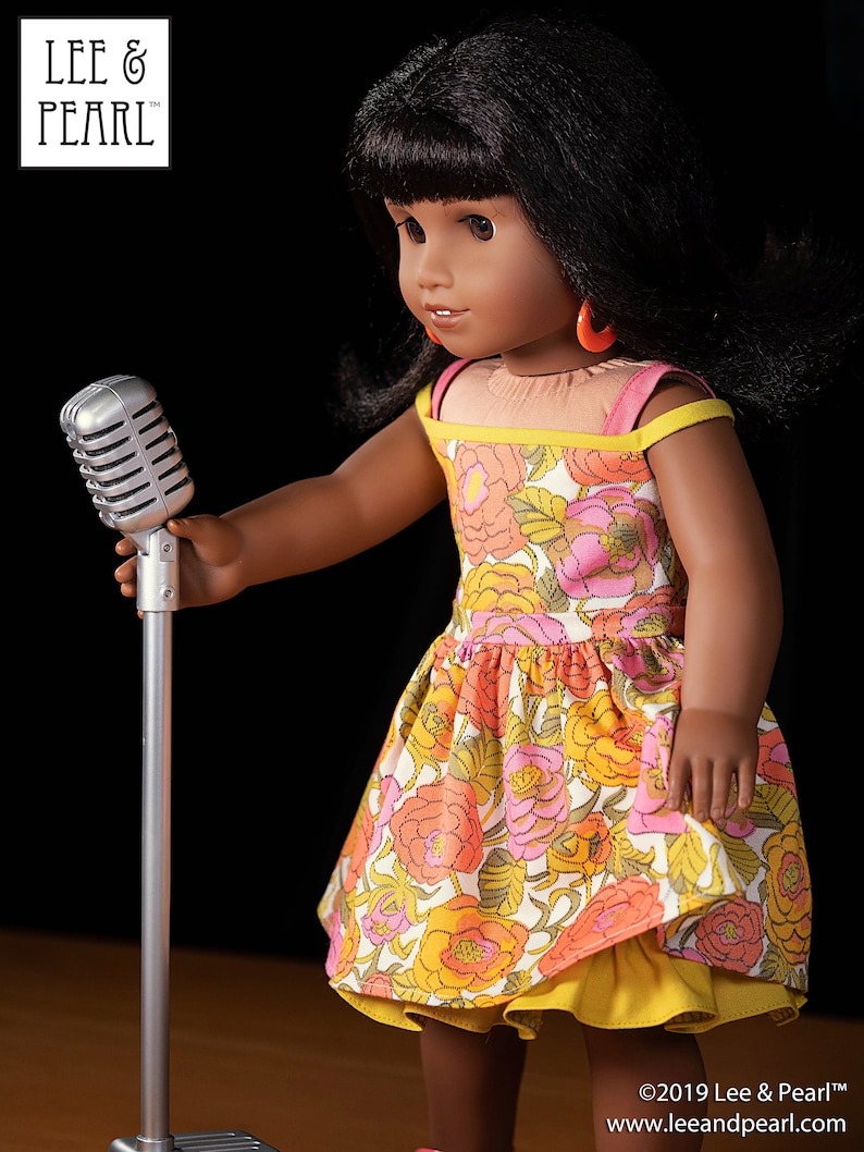 May include: A doll wearing a floral dress with a yellow underskirt and red earrings holds a silver microphone on a stand. The doll has dark hair and brown eyes. The background is black.