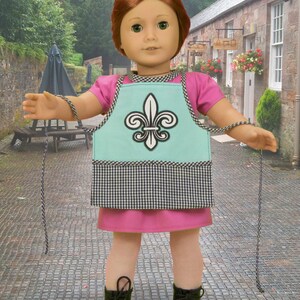 May include: A doll wearing a pink dress and a blue and white checkered apron with a fleur-de-lis design. The doll is holding a string in each hand.