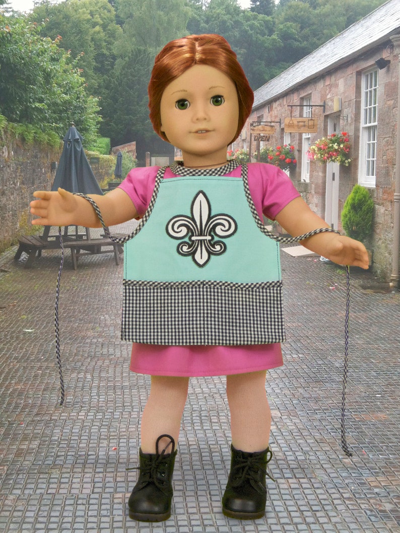 May include: A doll wearing a pink dress and a blue and white checkered apron with a fleur-de-lis design. The doll is holding a string in each hand.